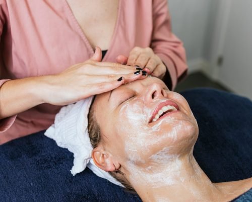 Crop unrecognizable cosmetician applying facial cleanser on face of female client during skin care treatment in beauty salon